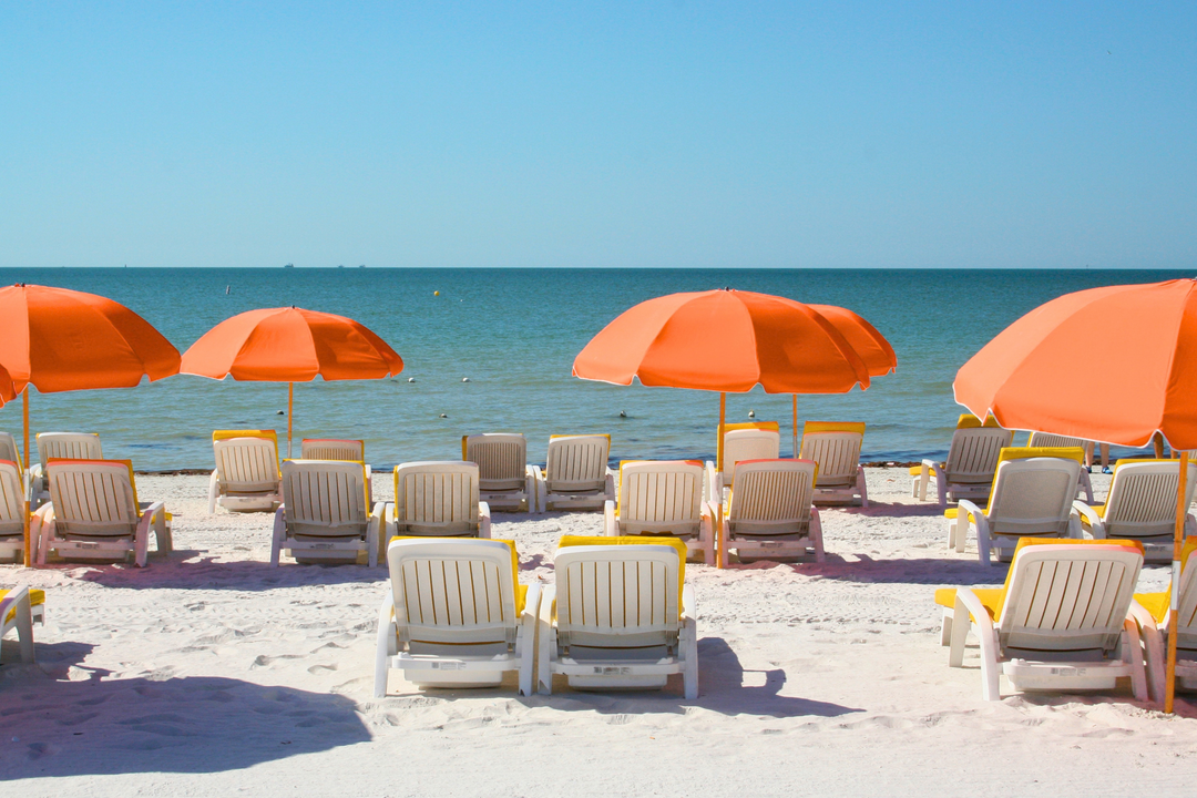 Hvide liggestole og orange parasoller på en sandstrand med udsigt over det blå hav.