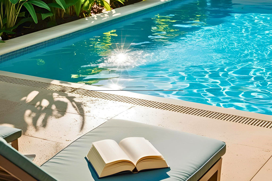 En åben bog ligger på en liggestol ved siden af en indbydende swimmingpool i solskinnet.
