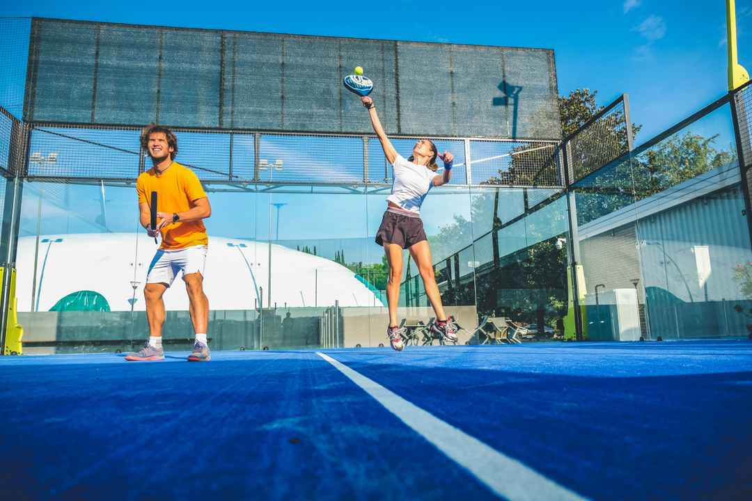 Et par spiller padeltennis på en blå udendørsbane under en skyfri himmel.